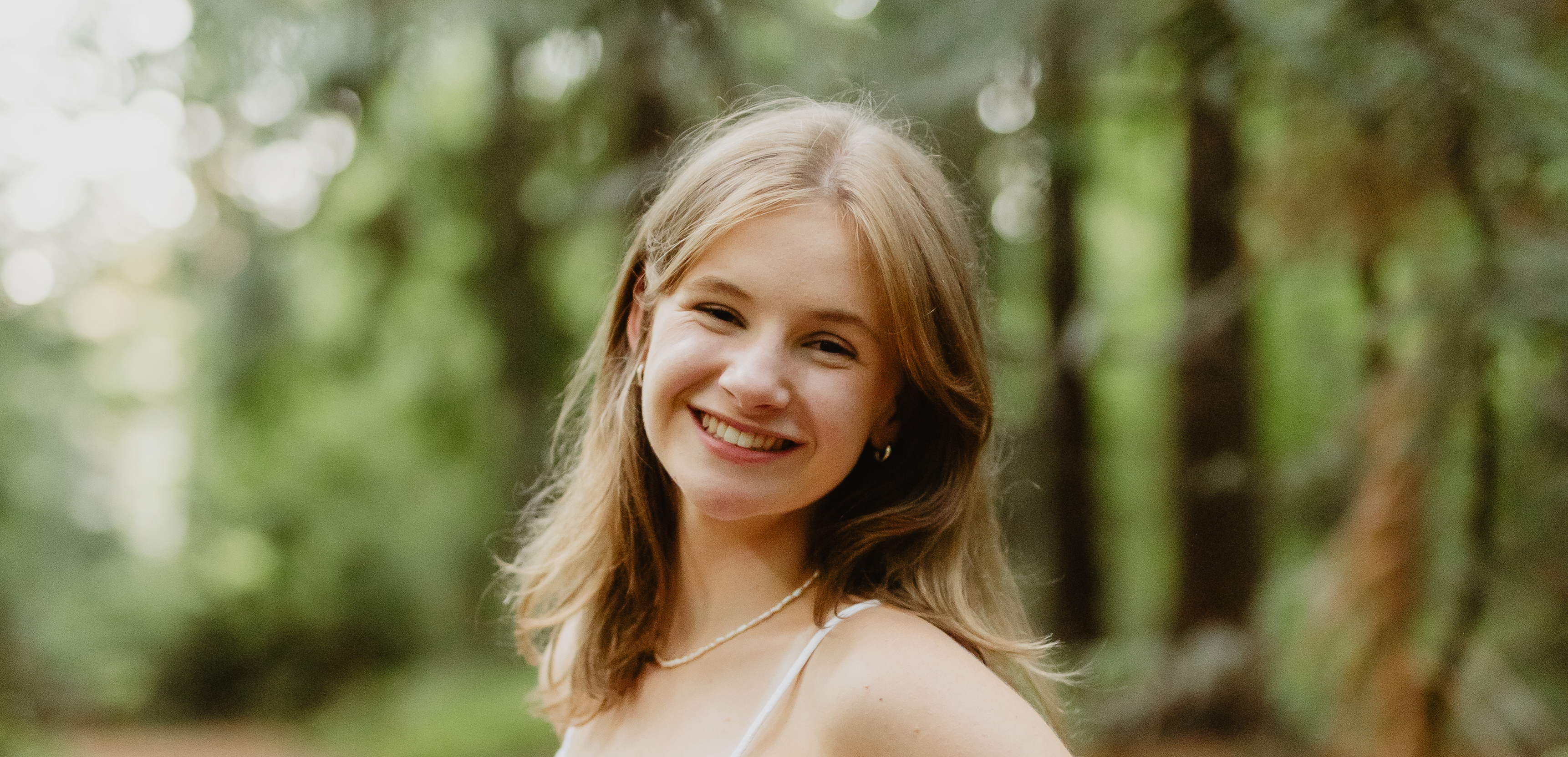 Portrait of a current Gannon Scholar with shoulder-length blonde hair, smiling, wearing a light-colored top and small hoop earrings, standing outdoors with trees softly blurred in the background.